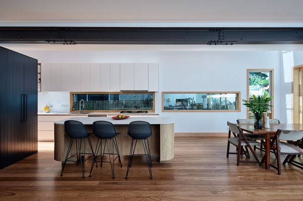 Open-plan contemporary kitchen and dining area with a large curved island, three bar stools, extensive white cabinetry, dark tall pantry cabinetry, timber flooring, and a glazed internal splashback or viewing panel. A dining table and chairs sit beside the kitchen, with greenery visible through the windows. A broad dark-framed opening or structural beam spans the ceiling.