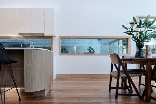 A bright, contemporary open-plan kitchen and dining area with warm timber flooring and cabinetry. The kitchen features a curved wood-grain island with a dark countertop, white upper cabinetry, and a blue-grey tiled splashback. A large interior pass-through or glazed opening with a timber frame reflects the kitchen, while a dark timber dining table and chairs occupy the right side of the room.