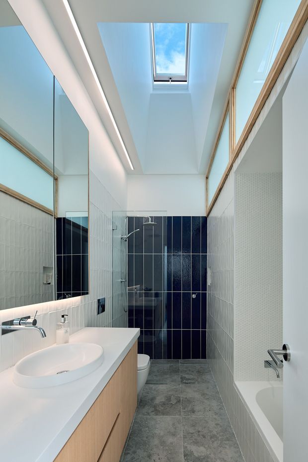 A bright, narrow contemporary bathroom with a skylight, full-height mirrors, white wall tiles, dark navy shower feature tiles, and grey stone-look floor tiles. A white vanity with integrated basin and light timber cabinetry runs along the left side, while a bathtub is partially visible on the right. Frameless glass shower screening, timber-framed glazing or mirrors, and recessed linear lighting create a clean, elongated space.