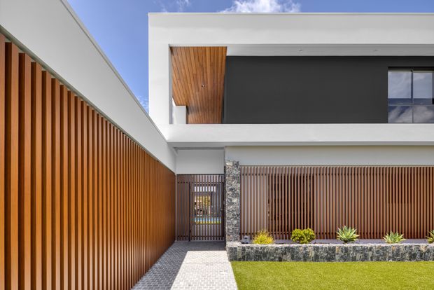 Contemporary residential facade viewed from the front approach, featuring crisp white rendered volumes, a dark charcoal upper wall, extensive warm timber vertical screening, a timber-lined recessed balcony or soffit, and a stone-clad entry column. A paved walkway leads past a low planted garden bed and lawn to a slatted pedestrian gate.
