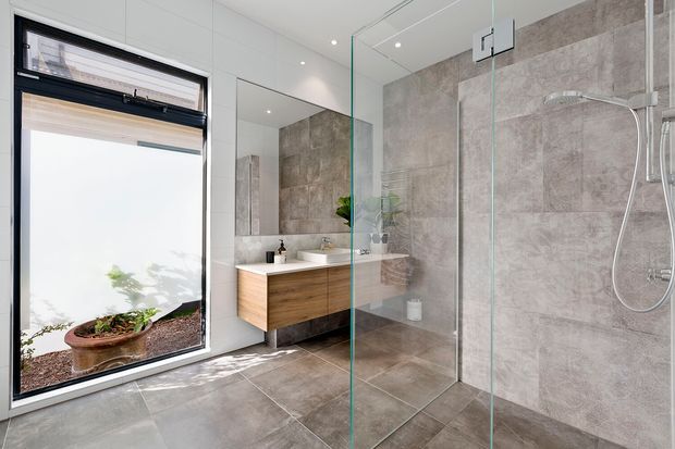 Bright contemporary bathroom with a frameless glass shower enclosure, large-format grey stone-look wall and floor tiles, a floating timber vanity with white countertop and basin, and a black-framed awning window overlooking a planted area. Recessed ceiling downlights and extensive mirrors enhance the clean, airy space.