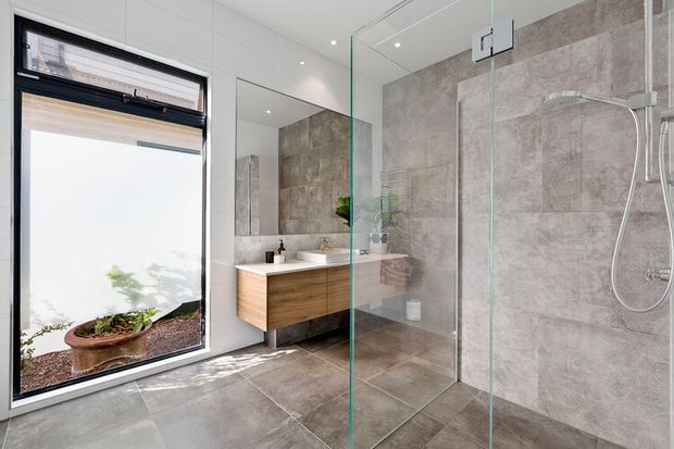 Bright contemporary bathroom with a frameless glass walk-in shower, large grey-beige tiled walls and floor, and a floating timber-veneer vanity with a white basin. A large black-framed window provides natural light and overlooks a small planted area; a wall mirror and recessed ceiling downlights complete the clean, modern fit-out.