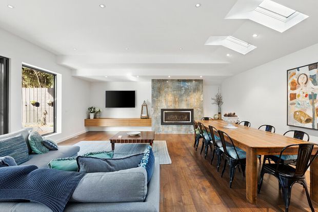 Open-plan contemporary living and dining area with a large blue sectional sofa in the foreground, a long timber dining table with black chairs, timber flooring, white walls and ceiling, skylights, recessed lighting, and a media wall with a wall-mounted television and low timber cabinetry. Artwork and decorative objects add visual interest, while large glazing brings in natural light.