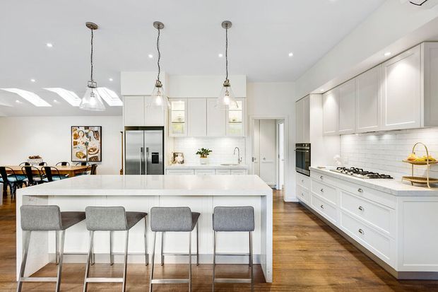 Bright, spacious open-plan kitchen with a large white island and four grey upholstered stools, extensive white shaker-style cabinetry, integrated appliances, a gas cooktop, pendant lights, and timber flooring. A dining area is visible beyond the island, with skylights and recessed ceiling lighting contributing to the airy feel.