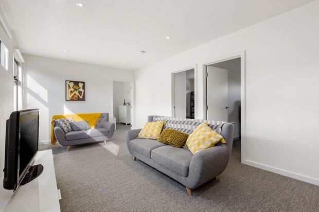 Bright contemporary living room with two grey upholstered sofas, patterned yellow and neutral cushions, a wall-mounted television, light grey carpet, white walls and ceiling, recessed downlights, and multiple doorways leading to adjacent rooms. Strong natural light enters through windows on the left.
