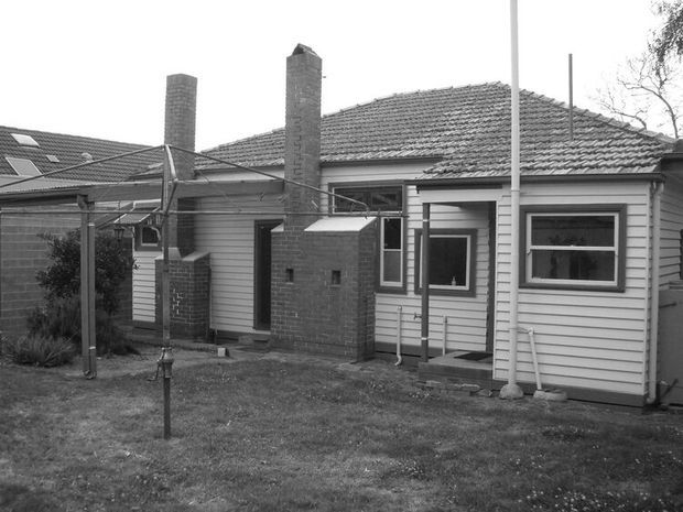 Black-and-white exterior photograph of a single-storey weatherboard house with a hipped tiled roof, multiple brick chimneys, and a small covered entry porch. A simple yard, clothesline structure, and neighbouring roof are visible. The image appears documentary or archival in style.