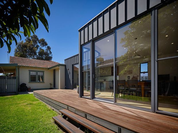 Exterior view of a contemporary dark-clad home extension with large floor-to-ceiling glazed doors opening onto a raised timber deck and stepped seating. A modern pavilion-like addition sits beside an older weatherboard house with a tiled pitched roof, creating a clear contrast between new and existing architecture. The scene includes a lawn and mature trees, with a bright blue sky and strong sunlight.