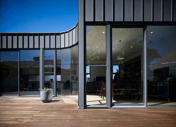 Contemporary single-storey building facade opening onto a broad timber deck. The image focuses on dark-framed floor-to-ceiling glazing, glazed doors, and a distinctive curved roofline or clerestory band with vertical dark metal framing. A dining and living area is visible through the glazing, while a large planter sits on the deck. Bright daylight and strong reflections create a clean, modern indoor-outdoor connection.