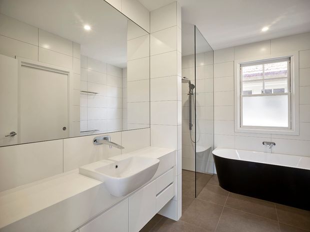 A bright, contemporary bathroom with a wall-mounted white vanity, vessel basin and chrome tapware. Large mirrored panels extend across the vanity wall, while a frameless glass shower enclosure sits centrally beside a freestanding black-and-white bathtub. The room has white tiled walls, warm grey-brown floor tiles, recessed ceiling lights and a frosted lower window.
