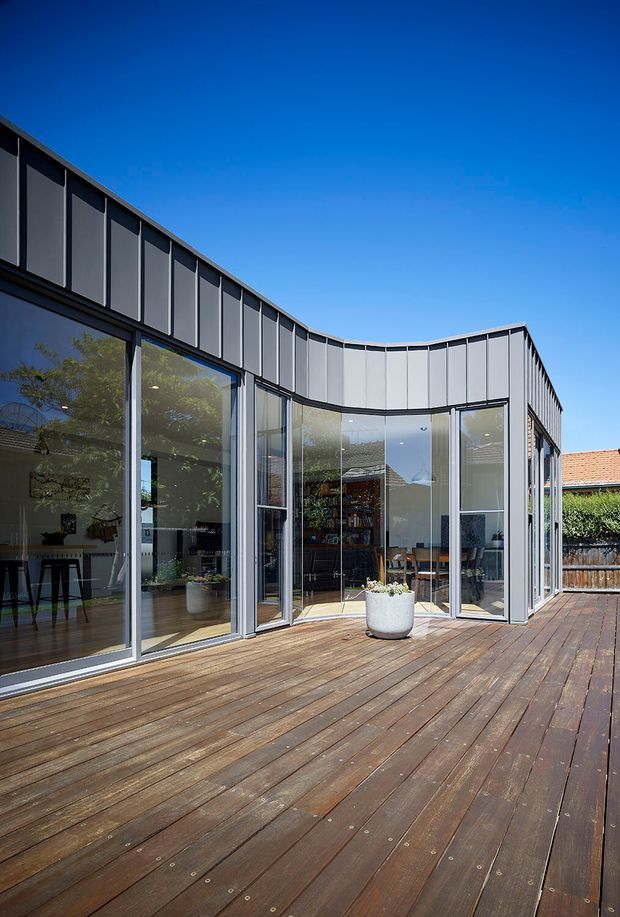 Exterior view of a contemporary curved-wing building opening onto a broad timber deck. The facade features extensive floor-to-ceiling glazing with slender dark metal frames and vertical grey cladding panels, while the interior living and dining areas are visible through the glass. Bright clear sky and neighbouring residential elements form the background.