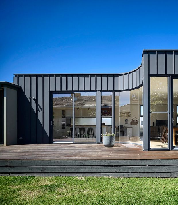Photograph of a contemporary dark-clad residential extension viewed from a timber deck and lawn. The facade combines standing-seam metal cladding with extensive floor-to-ceiling glazing and dark framed sliding or folding doors. The curved roof and wall profile forms a sculptural transition between the taller glazed volume and the lower adjoining section. A bright open-plan interior with kitchen island stools and dining furniture is visible through the glazing.