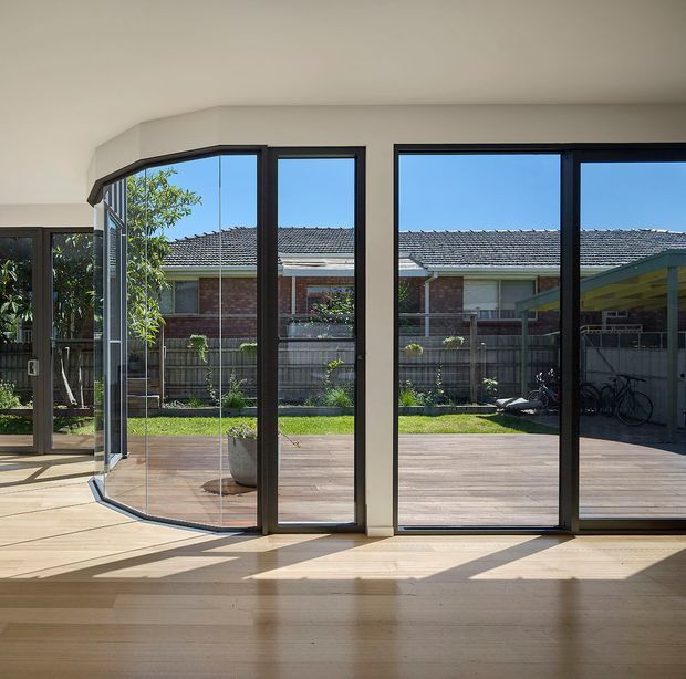 Bright contemporary interior opening onto a timber deck and landscaped backyard through expansive dark-framed floor-to-ceiling glazing. The view emphasizes a corner arrangement of large sliding or folding glass doors, pale timber flooring, strong sunlight, and a covered outdoor area with bicycles beyond.