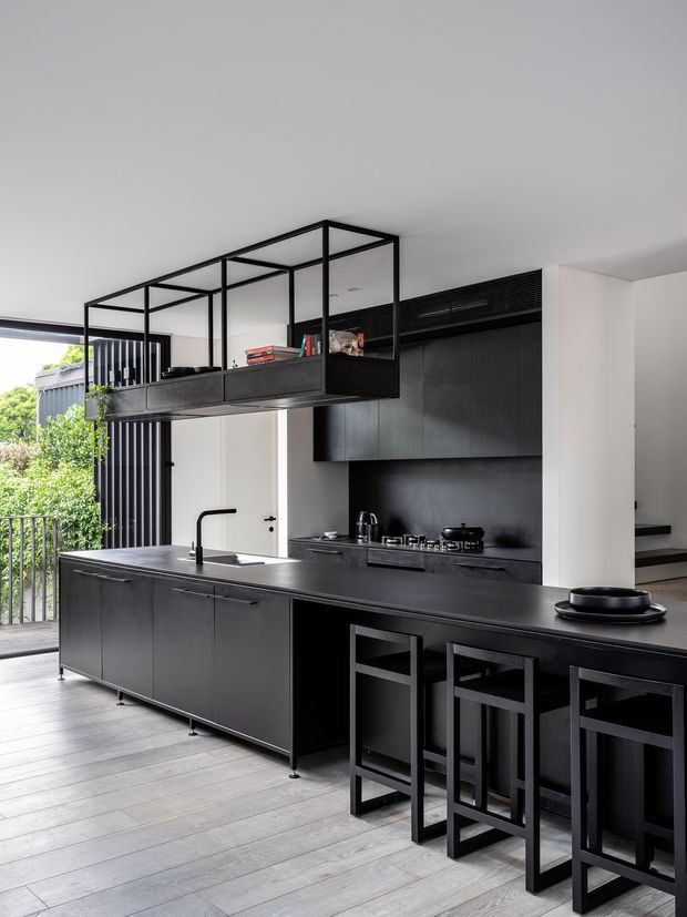 A sleek, dark-toned contemporary kitchen with a long island and integrated breakfast bar, black cabinetry, dark benchtops, and a matching dark splashback. A black metal-framed open shelving/mezzanine-style overhead structure adds architectural interest. Pale timber-look flooring, white walls, and large glazed openings create contrast and bring in natural light. Three black bar stools line the island, with greenery visible beyond the opening.