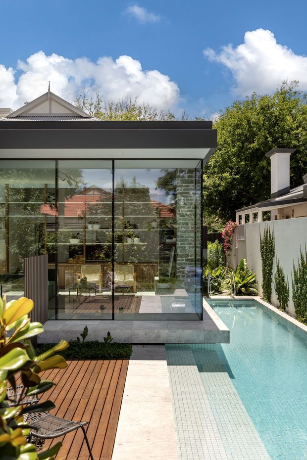 A bright exterior view of a contemporary glass-walled pavilion or extension opening onto a narrow tiled swimming pool. The structure has a flat dark roof, slim black-framed floor-to-ceiling glazing, and a light stone-clad base. A timber deck with outdoor chairs borders the pool, while lush planting and neighbouring residential roof forms are visible around the site.