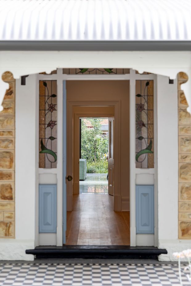 View through an ornate residential entryway into a sequence of interior rooms. The entrance has white painted architectural trim, pale blue lower panels, decorative stained-glass sidelights with floral motifs, sandstone surrounds, and a black threshold. A glossy black-and-white checkerboard tiled porch leads to a timber-floored hallway and open doorway beyond, with greenery visible at the far end.