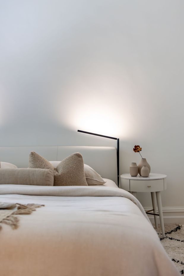 A softly styled contemporary bedroom in a neutral palette. The image shows a neatly made bed with layered cream and beige bedding and textured pillows, a padded light upholstered headboard, and a slim black linear wall-mounted reading light. A small round white bedside table holds ceramic vases and a dried floral stem. White walls, subtle skirting, and a plush neutral patterned rug create a calm, refined atmosphere.
