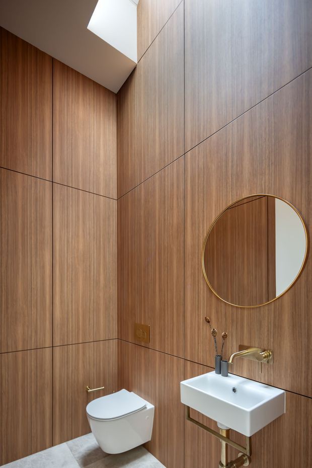 Compact contemporary bathroom with floor-to-ceiling warm wood-look wall panelling, dark linear grout joints, a wall-hung white toilet, and a floating rectangular white basin with exposed brass-toned plumbing. A round thin brass-framed mirror and angled ceiling detail add refined modern accents.