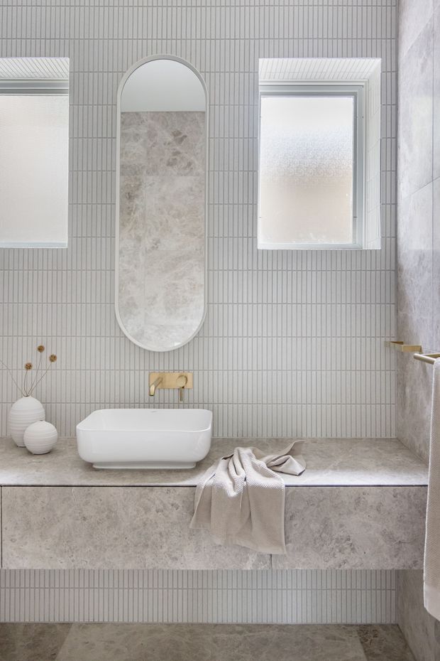 A bright, refined bathroom featuring a wall-mounted white vessel basin on a pale stone vanity, a built-in stone-clad bathtub, vertically ribbed light-grey wall tiles, matching stone-look floor and bath surround, an arched recessed mirror, frosted windows, and brushed brass tapware. Soft neutral styling and textured towels create a calm, contemporary atmosphere.