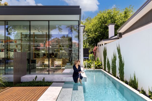 Contemporary residential courtyard with a narrow lap pool beside a glass-walled pavilion or living area. The composition features dark framed floor-to-ceiling glazing, a flat roof, pale rendered boundary walls, vertical planting, stone paving, and a timber deck under a bright blue sky.