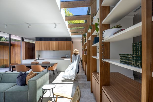A bright, open-plan contemporary interior combining a living and dining area with extensive custom timber shelving and joinery. The room features a pale sectional sofa, round dining table with upholstered chairs, timber wall panelling, white built-in shelving, a tiled or stone-look floor, and a glazed overhead timber pergola/skylight structure bringing in natural light. A woman in a white dress is present, but the focus is the residential interior and its built-in design.