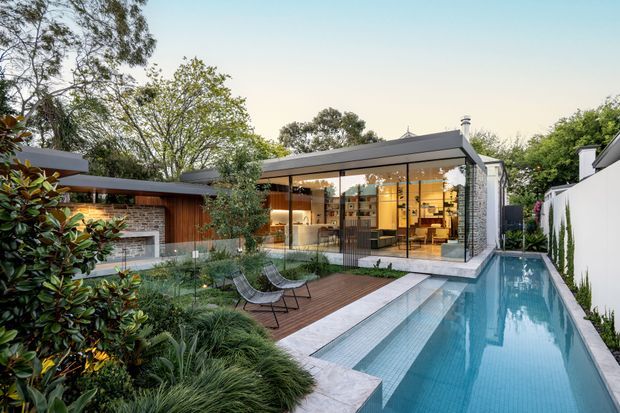 Exterior view of a contemporary single-storey residence with extensive floor-to-ceiling glazing, a flat roof, and a compact linear swimming pool. The landscaped courtyard includes a timber deck, outdoor lounge chairs, and a covered outdoor fireplace pavilion, while warm-lit interiors are visible through the glazing.