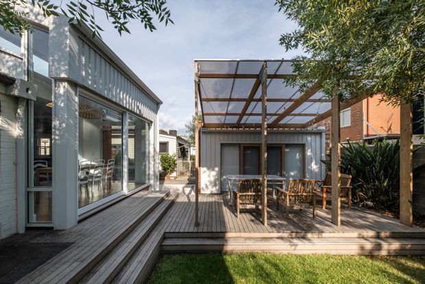 A contemporary residential courtyard viewed from the garden, featuring a raised timber deck connecting a glazed extension on the left with a compact grey-clad pavilion at the rear. A timber pergola with translucent roofing shelters an outdoor dining area, while established planting and lawn frame the space. The composition highlights indoor-outdoor connection, abundant glazing, timber decking and a private urban garden setting.