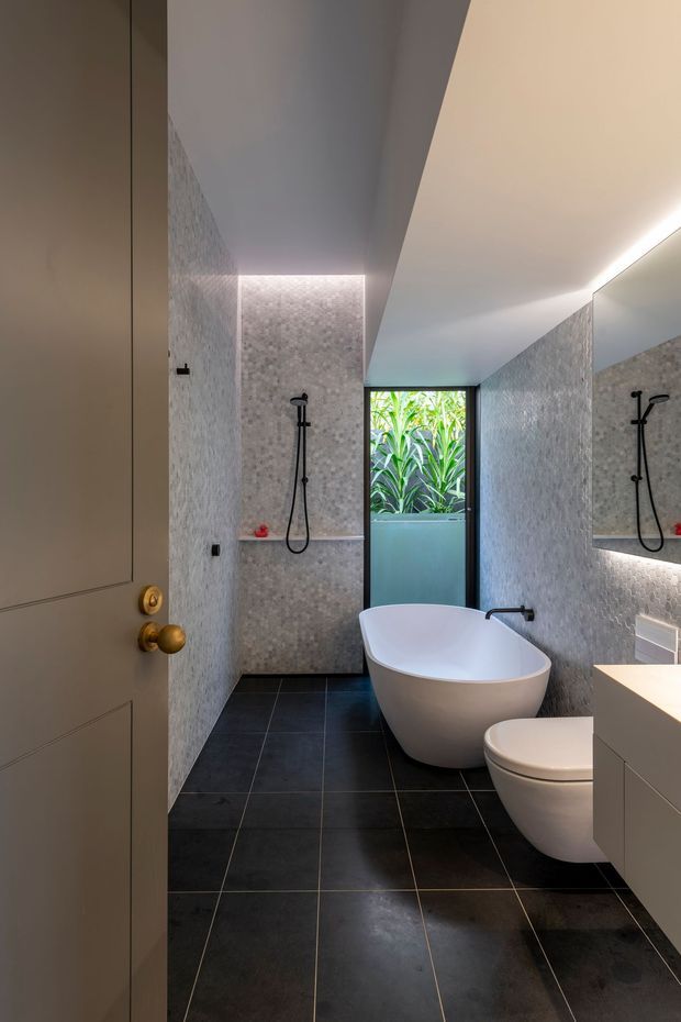 A contemporary bathroom viewed through an open door, featuring pale textured wall tiles, large dark floor tiles, a freestanding white oval bathtub, wall-mounted black shower fittings, a toilet, and a floating white vanity. A large illuminated mirror and concealed linear lighting add a clean, refined feel, while a frosted-glass door/window brings in greenery.