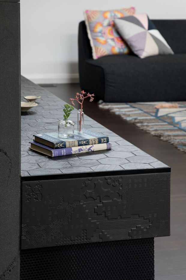 Close-up of a contemporary living room featuring a large black sofa with colourful patterned cushions, a dark decorative built-in console or coffee table with a textured geometric front, and a grey irregular-tile tabletop. Books and small glass vases with branches sit on the surface, while a patterned rug and dark flooring are visible in the background.