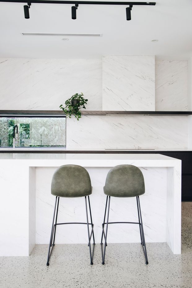 Bright contemporary kitchen featuring a large white marble-look island with integrated seating, matching full-height splashback and cabinetry, dark horizontal shelving, black track lighting, a stainless sink by a wide window, and two green upholstered bar stools. The floor is light terrazzo with dark aggregate, and a small potted plant adds greenery.