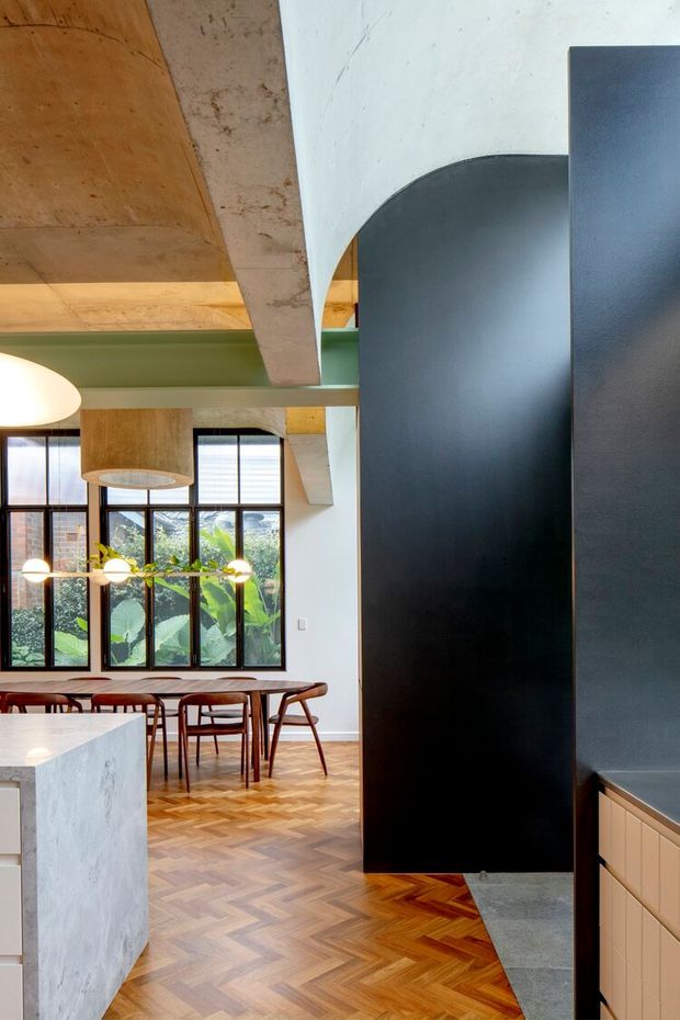A refined open-plan kitchen and dining space with a dramatic black curved kitchen volume, pale stone island, light timber cabinetry, and warm herringbone timber flooring. The dining area features a timber table and chairs beneath globe pendant lighting, with large black-framed windows overlooking lush greenery. Exposed concrete beams and timber ceiling elements contrast with a muted green structural accent.