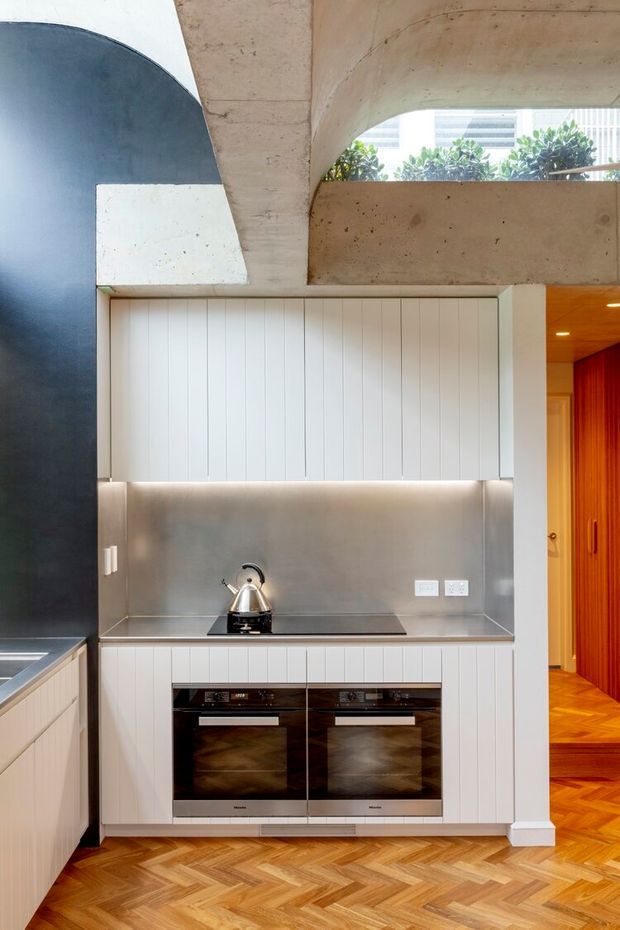 Compact contemporary kitchen set within a concrete architectural interior. The space features white vertically panelled cabinetry, a stainless-steel splashback and benchtop, integrated black ovens, and warm herringbone timber flooring. A dark curved wall and exposed concrete arch add strong sculptural and industrial character, with a brightly coloured timber-lined hallway visible beyond.