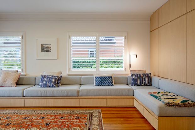 Warm, light-filled living room with a large built-in L-shaped upholstered sectional and integrated timber storage cabinetry. White plantation shutters cover the windows, while a patterned rug and blue-toned cushions add colour against the pale neutral walls and warm timber floor. A timber-paneled built-in wall rises to the ceiling on the right.