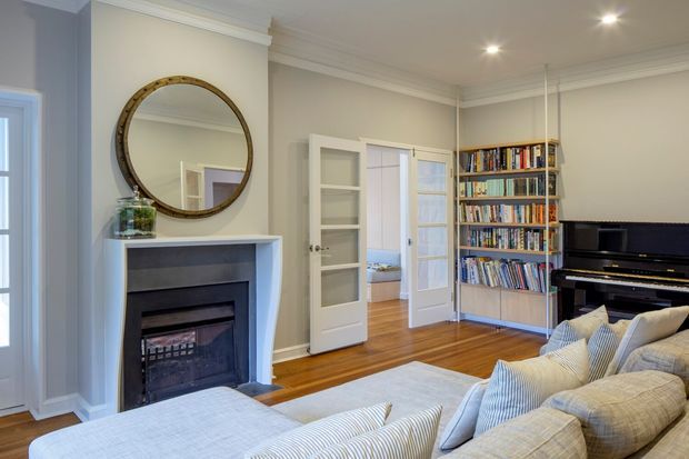 Bright, traditional-style living room with a white fireplace surround and dark inset firebox, round timber-framed mirror, pale walls with decorative cornices, polished timber flooring, glazed internal French doors, built-in timber shelving filled with books, and a black upright piano. A large pale sectional sofa occupies the foreground.
