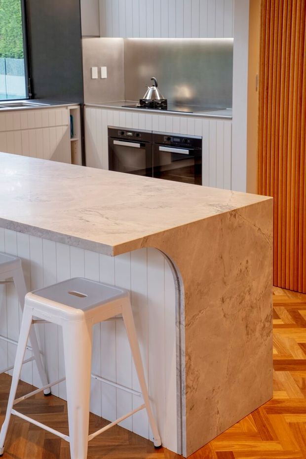 A compact, contemporary kitchen with a large pale stone island featuring a waterfall-style side panel and curved cut-out detail. White vertical wall panelling and cabinetry surround integrated black ovens, while a stainless-steel backsplash and benchtop run beneath a window. A white metal bar stool sits at the island, and a warm timber slatted screen and parquet timber flooring add contrast.