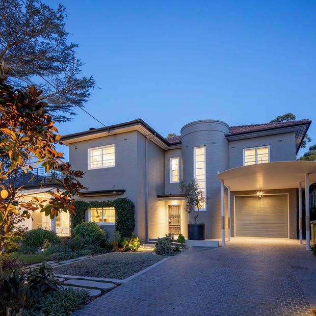 Exterior twilight view of a substantial two-storey residence with pale rendered masonry walls, a red tiled pitched roof, prominent cylindrical turret-like bay, illuminated windows, and an attached covered carport. The landscaped front garden includes clipped hedges, planting beds and gravel areas, while the driveway is finished in dark interlocking pavers.