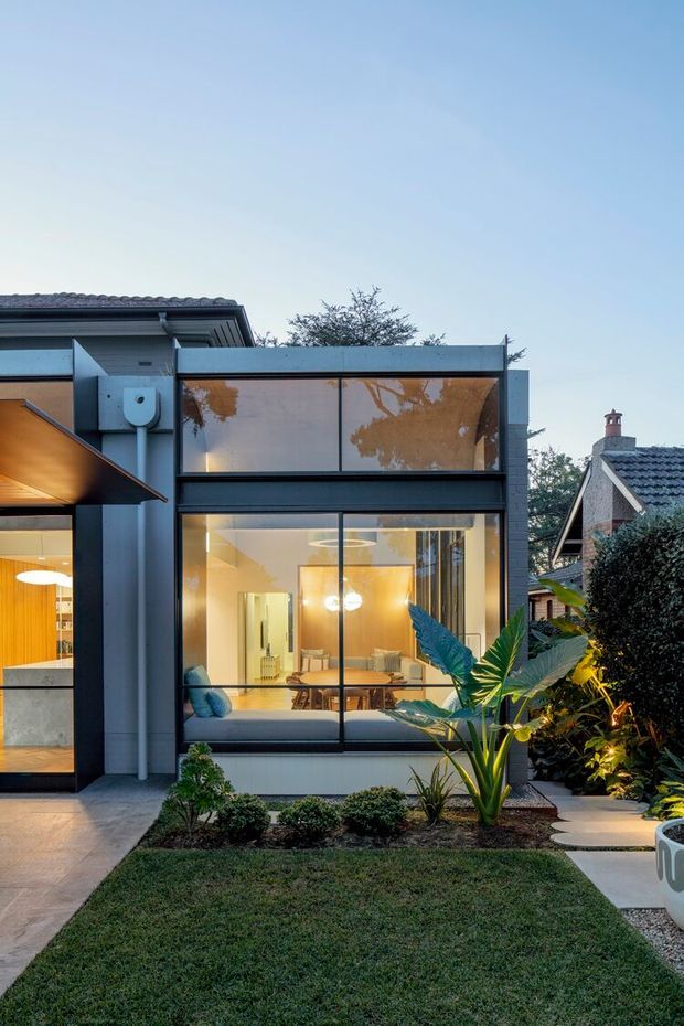 A contemporary residential rear extension viewed from a landscaped garden at dusk. The addition features dark metal-framed floor-to-ceiling glazing, a projecting covered outdoor area, and warm interior lighting visible through the windows. A small lawn, paved path, and tropical planting sit in the foreground, with the original tiled-roof house visible on either side.