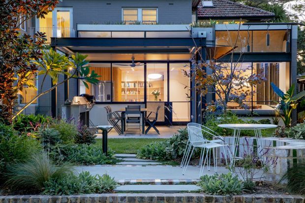 A modern multi-level residential extension viewed from a landscaped garden at dusk. The dark-clad facade has extensive floor-to-ceiling glazing, a projecting upper balcony, and a tiled pitched roof over part of the building. Warm interior lighting reveals an open-plan dining and living area, while the garden includes paved stepping paths, lush planting, an outdoor dining setting, and a built-in outdoor kitchen or barbecue zone.