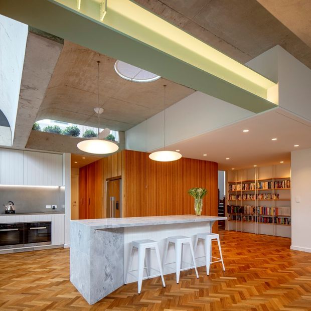 Open-plan kitchen and living area with a large pale stone island, white cabinetry, integrated appliances, timber-paneled feature wall, exposed concrete ceiling, and built-in bookshelf. The floor is laid in a warm herringbone timber pattern, with pendant lights above the island and a visible skylight.
