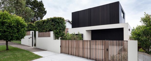 Contemporary two-storey residential facade with a strong geometric composition. The upper level is clad in dark vertical ribbed panels, contrasting with crisp white rendered walls and a recessed dark garage opening. A vertical timber slat fence and gate screen the front garden and driveway, with mature landscaping around the property.
