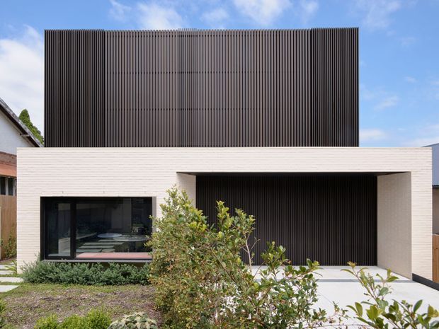 Street-facing exterior of a contemporary two-storey residence. The lower level is clad in pale white brick with a broad recessed carport or covered entry, while the upper storey is screened with closely spaced dark vertical battens. Dark-framed glazing and restrained landscaping complete the modern facade.