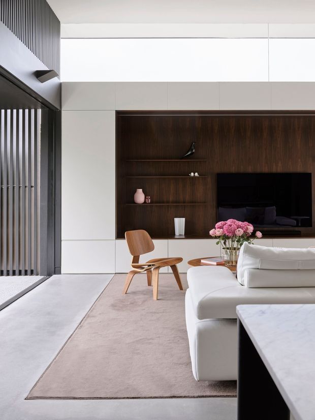 Contemporary living room with a white sectional sofa, pale area rug, sculptural wooden lounge chair, and a dark timber built-in media wall with open shelving and television. Large black-framed glazing and vertical screening provide strong architectural contrast, while pale wall panels and a marble-topped table create a refined, minimalist composition.