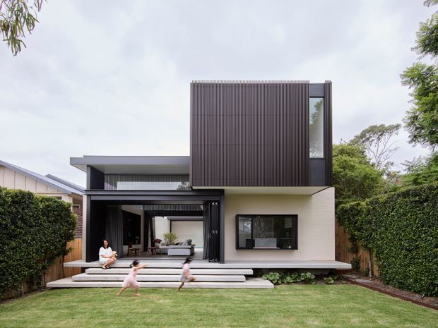 Contemporary two-storey residential house viewed from the rear garden. The home has a strong rectilinear composition, a projecting upper-level volume clad in dark narrow vertical battens, pale rendered walls, broad glazing, and a covered outdoor living area opening onto stepped concrete terraces and a lawn. Dense hedging and mature trees frame the suburban setting.