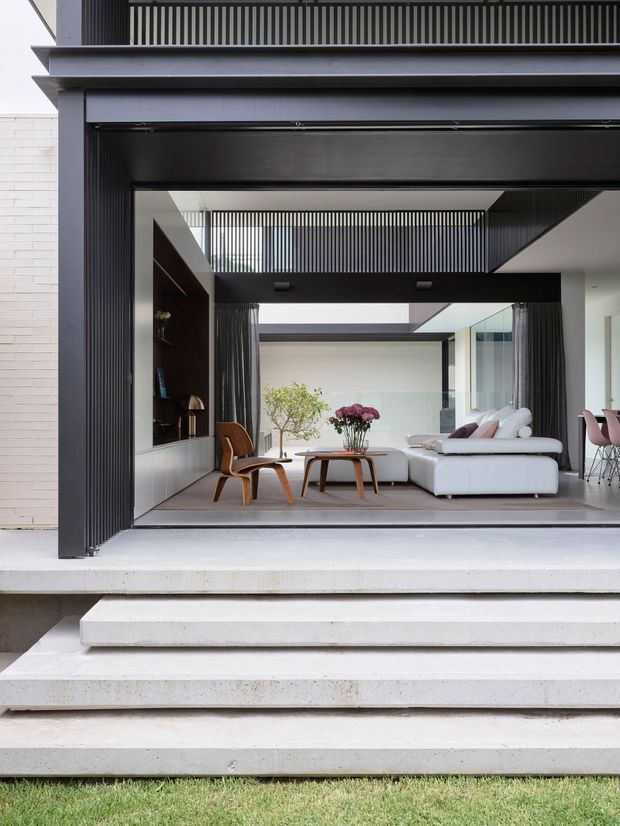 A contemporary residential indoor-outdoor living space viewed from the garden, framed by a large dark charcoal opening with vertical screening and a broad overhead structure. Wide pale concrete steps lead up to a sunken or raised living area furnished with a white sectional sofa, timber lounge chair, low timber coffee table, and flowers. Glazing, curtains, a dark built-in shelving unit, and glimpses of a dining area are visible inside. The composition emphasises strong indoor-outdoor connection, geometric lines, and contrasting dark architectural framing with pale concrete and white furnishings.