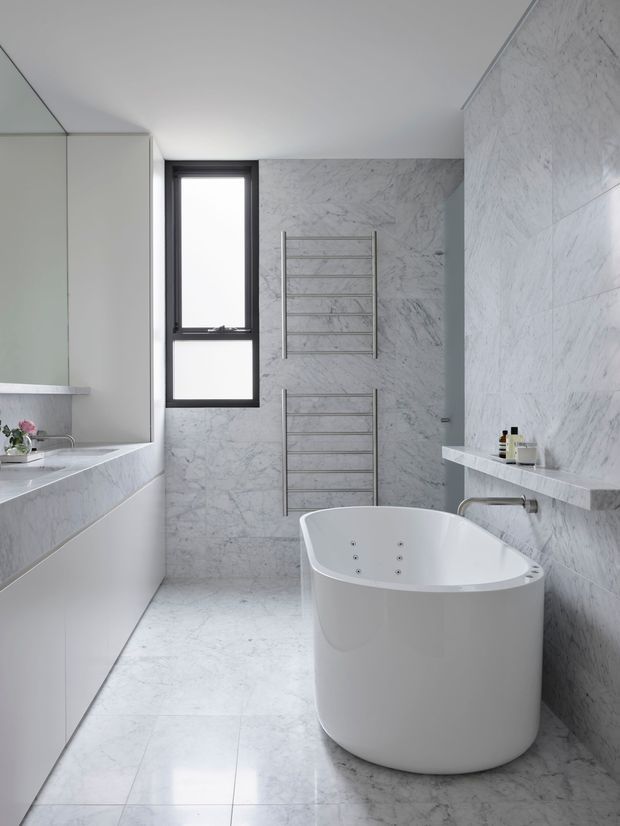 A bright, contemporary bathroom clad in white-grey veined marble-look surfaces. The room features a freestanding white oval bathtub, wall-mounted chrome tapware, two chrome heated towel rails, a large mirrored vanity with integrated basin, and a narrow black-framed window. The clean, pared-back design has strong tonal consistency and polished modern fittings.