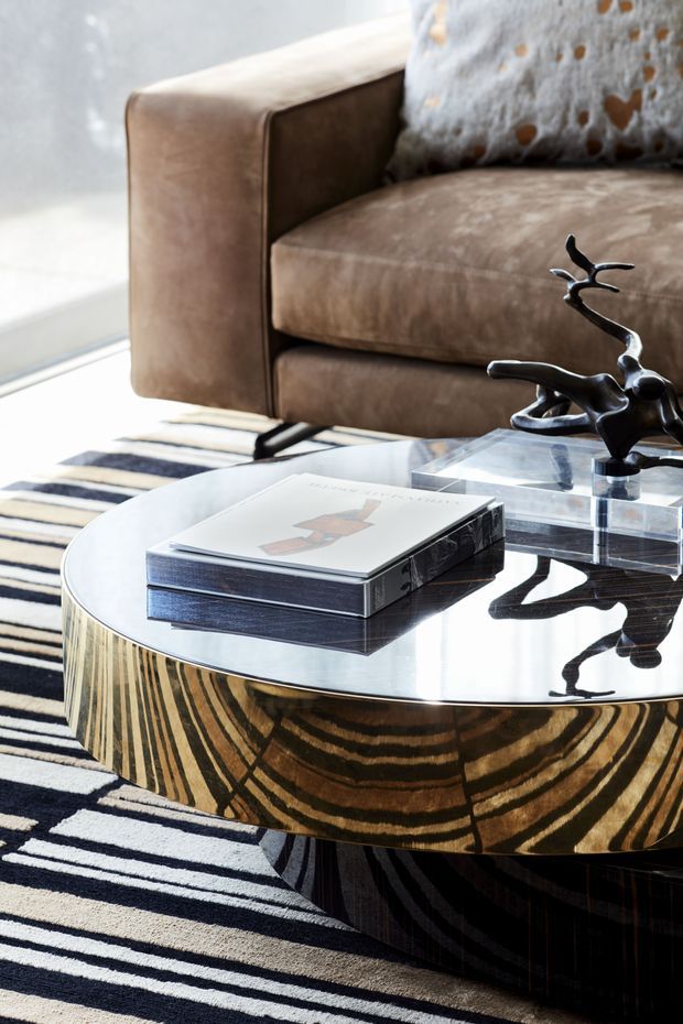 Close-up interior view of a refined living room seating area. A brown upholstered armchair sits behind a round coffee table with a reflective glass top and a dramatic striped wood-grain base, accented by a sculptural dark metal object and stacked books. A bold black-and-cream striped rug and decorative cushion complete the layered, high-contrast styling.