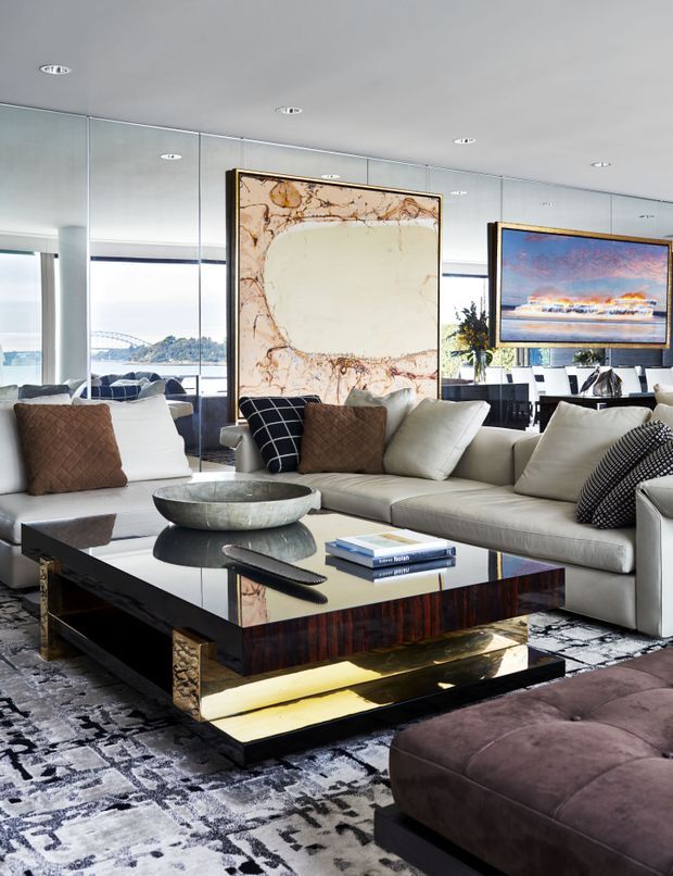 Luxurious contemporary living room with a large cream sectional sofa, mirrored wall panels, oversized framed artwork, a reflective dark wood and metallic coffee table, patterned area rug, and plush ottoman. Floor-to-ceiling glazing reveals a bright waterfront outlook beyond the room.