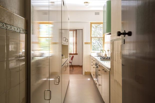 A narrow, bright galley kitchen viewed from an open doorway. Full-height cream cabinetry lines the left side, while the right side has lower cabinets, a light benchtop, cooktop and sink beneath timber-framed windows. Cream tiled walls feature a decorative horizontal border, with warm timber trim and a muted green upper cabinet adding colour. A small dining or sitting area is visible at the far end.