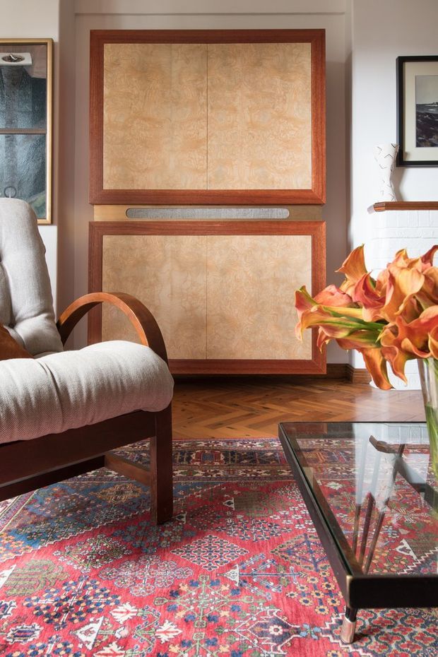 A warmly styled living room corner featuring a vintage upholstered wooden armchair, richly patterned Persian-style rug, glass coffee table, framed artwork, a white brick fireplace edge, and prominent custom wall panelling with reddish timber frames and textured tan inset panels. A vase of orange flowers adds a vivid foreground accent.