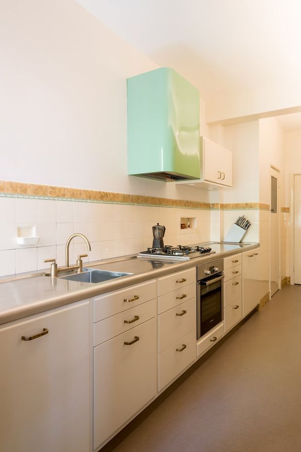 A compact, galley-style kitchen with white built-in cabinetry, stainless-steel benchtops and integrated appliances. The room features a tiled backsplash with a decorative border, a double sink, gas cooktop, oven, pale green curved rangehood, and warm neutral flooring. The overall appearance is functional and retro-influenced with bright, clean finishes.