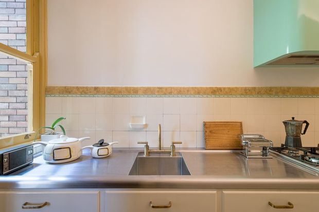 A compact retro-style kitchen viewed straight on, with a stainless-steel benchtop, integrated sink, pale painted cabinetry, tiled splashback with a decorative border, timber-framed window, and mint-green upper cabinetry. Small countertop appliances and cooking equipment sit along the bench.